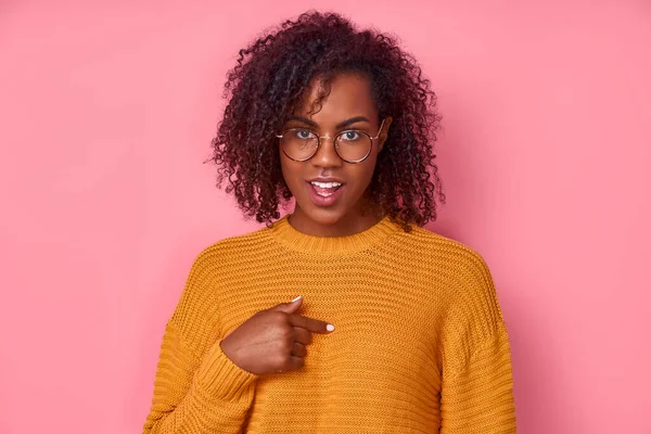 Słodki pozytywny młody Afroamerykanin kobieta w żółtej koszuli z okularami wskazuje na siebie, pyta o coś, ma szczęśliwy wyraz wyizolowany na różowym tle. Pojęcie świadomego wyboru — Zdjęcie stockowe