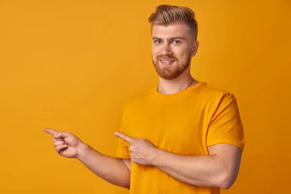 Glada ung man med skägg visar vänstra hörnet med båda pekfingrarna, ler med godkännande, föreslår att gå dit, på gul vägg. Begreppet reklam för bilverkstad för hushållsapparater — Stockfoto