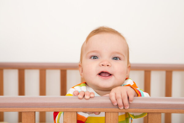 A baby is smiling in the crib and holds onto the side of the bed