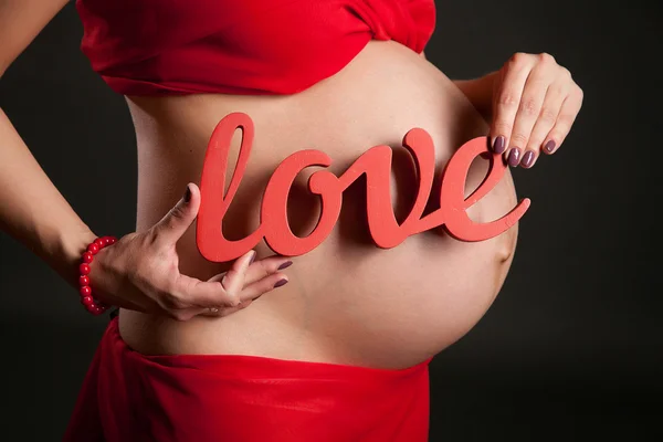 Ventre d'une femme enceinte dans une robe rouge, les mains de la mère avec le mot amour — Photo