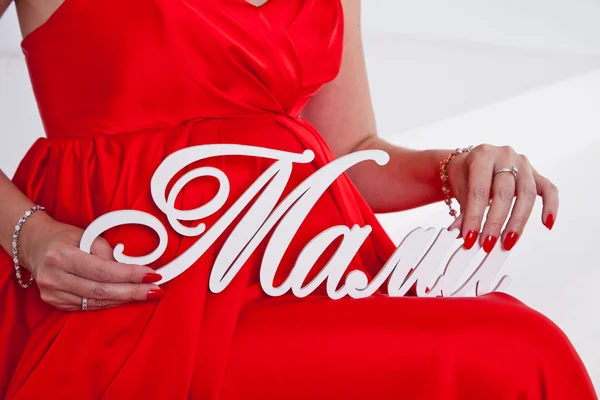 Barriga de uma mulher grávida em um vestido vermelho, mãos mãe segurando a palavra mama — Fotografia de Stock