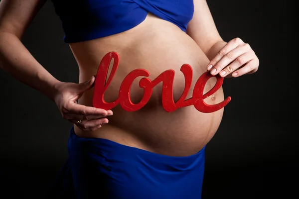 Belly of a pregnant woman in a blue dress, mother hands holding the word love — Stock Photo, Image