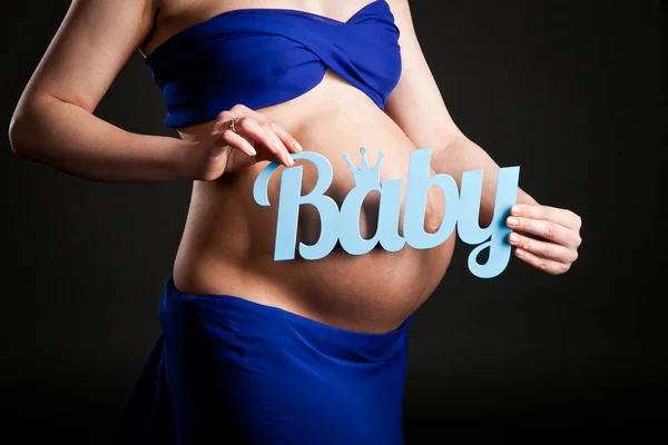 Belly of a pregnant woman in a blue dress, mother hands holding the word baby — Stock Photo, Image