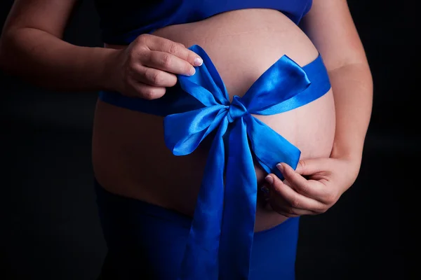 Ventre d'une femme enceinte dans une robe bleue, les mains de la mère avec arc bleu — Photo