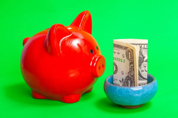 red pig piggy bank in front of a  bowl with money on a green background
