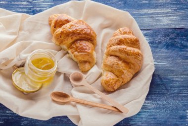 Çok güzel çıtır kruvasan ve hafif kumaş üzerinde bir kavanoz bal, ballı lezzetli pastalar ve mavi arka planda kumaş peçetede kaşıklar.