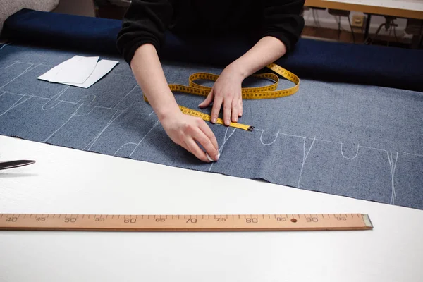 Manos de costurera cortando un tejido vaquero con patrón de tijeras y cinta métrica en mesa blanca —  Fotos de Stock