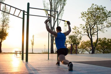 egzersiz süspansiyon ile açık spor salonunda, güçlü adam park, gündoğumu veya günbatımı deniz arka planda sabah erken eğitim kayışlar 