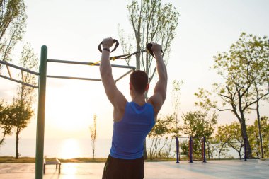 egzersiz süspansiyon ile açık spor salonunda, güçlü adam park, gündoğumu veya günbatımı deniz arka planda sabah erken eğitim kayışlar 