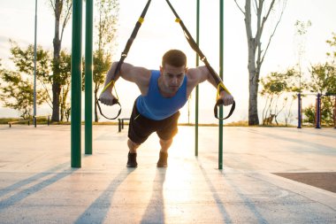 egzersiz süspansiyon ile açık spor salonunda, güçlü adam park, gündoğumu veya günbatımı deniz arka planda sabah erken eğitim kayışlar 