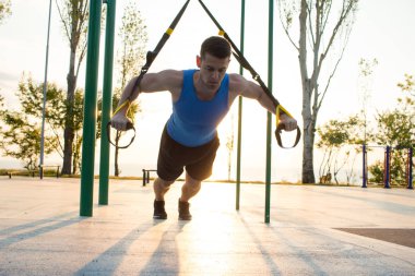 egzersiz süspansiyon ile açık spor salonunda, güçlü adam park, gündoğumu veya günbatımı deniz arka planda sabah erken eğitim kayışlar 