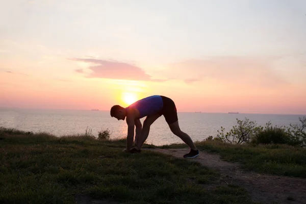 Giovane maschio che si allunga prima della sessione di allenamento al parco. Giovane sano che si riscalda all'aperto. Tramonto o alba in mare o ocen sullo sfondo — Foto Stock