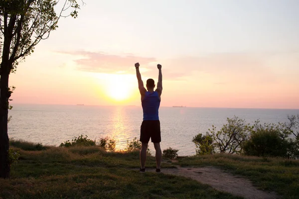 Corredor feliz viendo amanecer o atardecer con puños levantados, joven atleta sobre hierba durante el amanecer en el mar — Foto de Stock