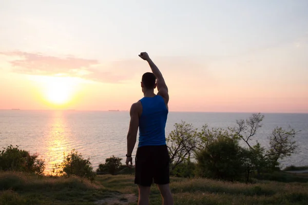 Corredor feliz viendo amanecer o atardecer con puños levantados, joven atleta sobre hierba durante el amanecer en el mar — Foto de Stock