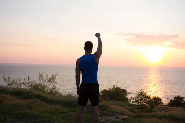 Happy fit corredor assistindo nascer do sol ou pôr do sol com punhos levantados, jovem atleta na grama durante o nascer do sol no mar — Fotografia de Stock