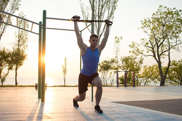 Egzersiz süspansiyon ile açık spor salonunda, güçlü adam park, gündoğumu veya günbatımı deniz arka planda sabah erken eğitim kayışlar — Stok fotoğraf