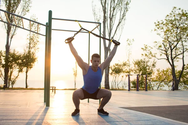 Egzersiz süspansiyon ile açık spor salonunda, güçlü adam park, gündoğumu veya günbatımı deniz arka planda sabah erken eğitim kayışlar — Stok fotoğraf