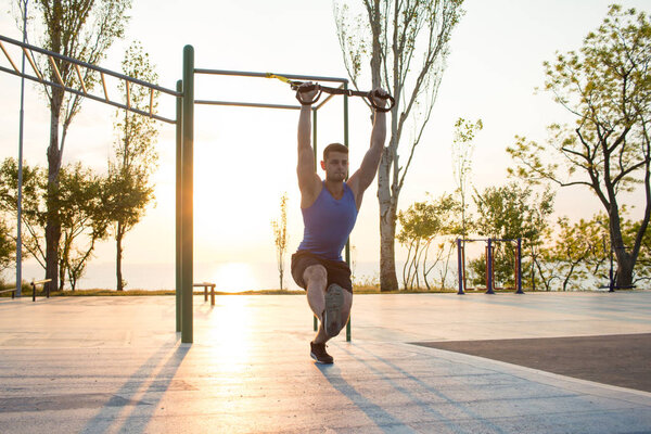workout with suspension straps In the outdoor gym, strong man training early in morning on the park, sunrise or sunset in the sea background 