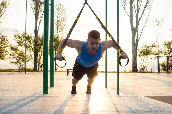 Egzersiz süspansiyon ile açık spor salonunda, güçlü adam park, gündoğumu veya günbatımı deniz arka planda sabah erken eğitim kayışlar — Stok fotoğraf