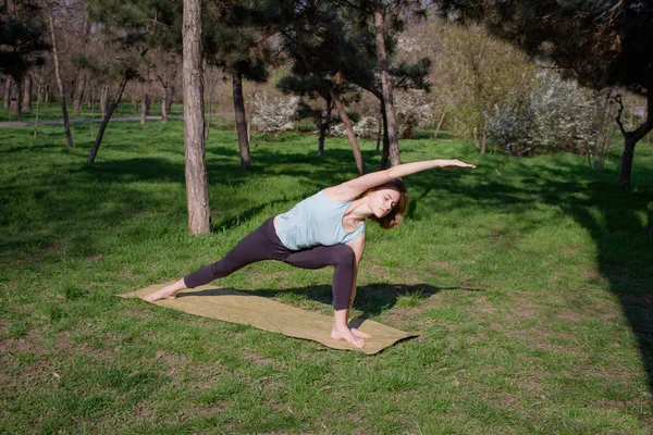 Giovane bella donna in forma facendo asans yoga nel parco dei pini con tappetino yoga — Foto Stock