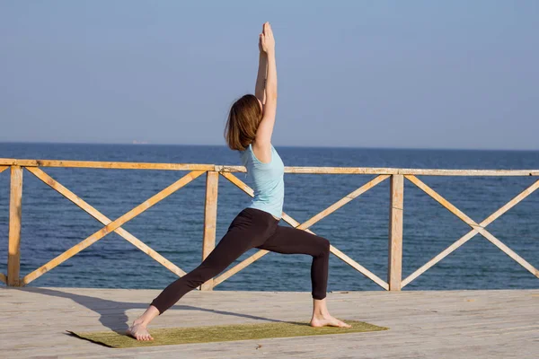 Mujer sexy joven haciendo ejercicios de yoga en el muelle de madera contra el fondo del mar y el cielo azul —  Fotos de Stock