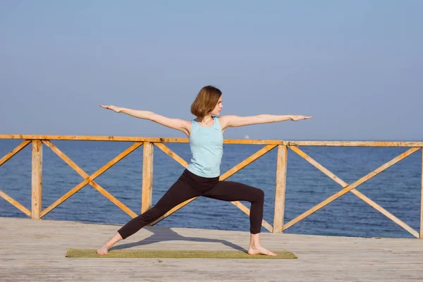 Jonge sexy vrouw doen yoga oefeningen op de houten pier tegen zee achtergrond en blauwe hemel — Gratis stockfoto
