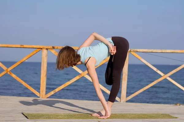 Mujer sexy joven haciendo ejercicios de yoga en el muelle de madera contra el fondo del mar y el cielo azul —  Fotos de Stock