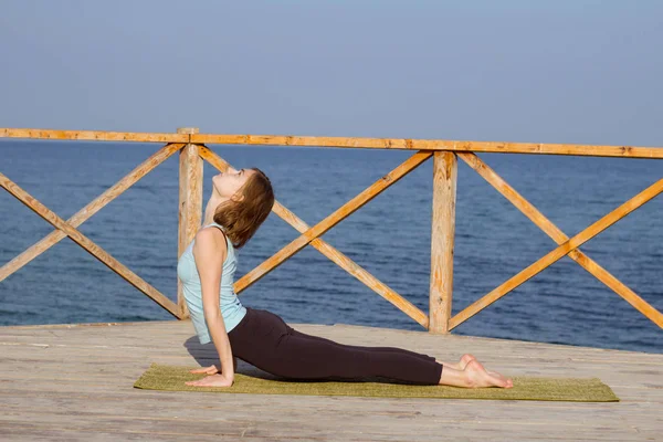 Tânăra femeie sexy face exerciții de yoga pe cheiul de lemn pe fundalul mării și cerul albastru — Fotografie, imagine de stoc