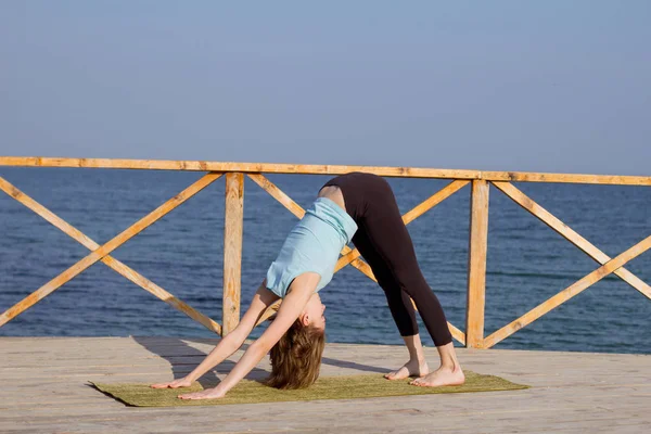 Mujer sexy joven haciendo ejercicios de yoga en el muelle de madera contra el fondo del mar y el cielo azul —  Fotos de Stock