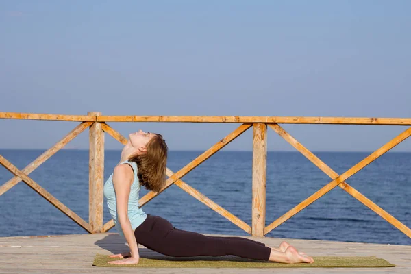 Genç seksi kadının deniz arka plan ve mavi gökyüzü karşı ahşap iskele üzerinde yoga egzersizleri yapması — Stok fotoğraf