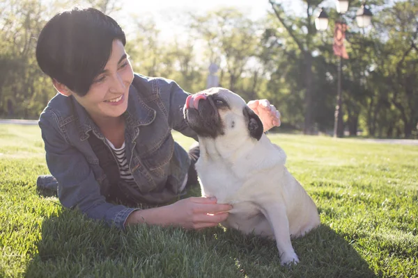 Porträt einer jungen Frau und eines Mops, die bei Sonnenuntergang oder Sonnenaufgang Zeit im Park auf grünem Gras verbringen — Stockfoto