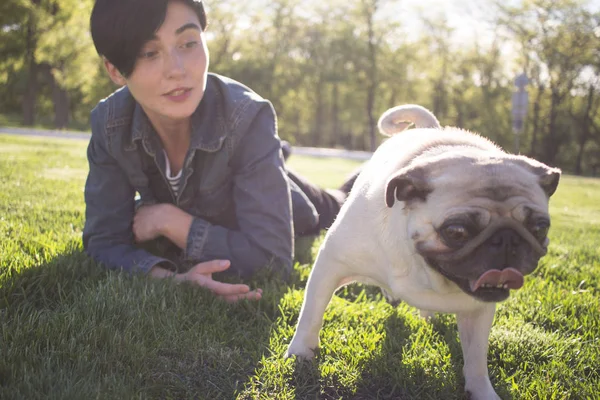 Retrato de jovem e pug tendo deus tempo no parque em grama verde durante o pôr do sol ou nascer do sol — Fotografia de Stock