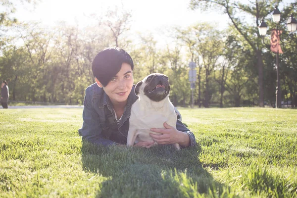 Retrato de jovem e pug tendo deus tempo no parque em grama verde durante o pôr do sol ou nascer do sol — Fotografia de Stock