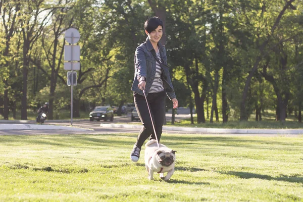 Junge Frau mit Mops im grünen Gras, hübsches Mädchen mit Hund beim Spielen im Park bei Sonnenuntergang oder Sonnenaufgang — Stockfoto