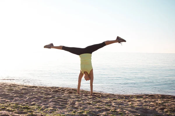 Молодая профессиональная гимнастка танцует на пляже, тренировки стоя на руках, восход солнца на море или в океане — стоковое фото
