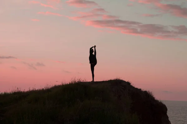 Passar kvinna som gör yoga stretching Motion utomhus i vackra landskap. Kvinna på vagga med havet och soluppgång eller solnedgång bakgrund utbildning asans. Siluett av kvinna i yogaställningarna — Stockfoto