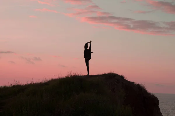 Passar kvinna som gör yoga stretching Motion utomhus i vackra landskap. Kvinna på vagga med havet och soluppgång eller solnedgång bakgrund utbildning asans. Siluett av kvinna i yogaställningarna — Stockfoto