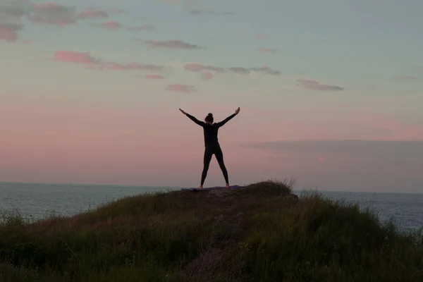 Passar kvinna som gör yoga stretching Motion utomhus i vackra landskap. Kvinna på vagga med havet och soluppgång eller solnedgång bakgrund utbildning asans. Siluett av kvinna i yogaställningarna — Stockfoto