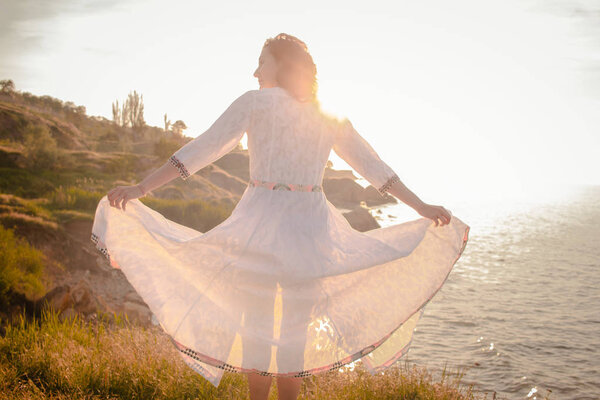 Young woman walking on the morning beach in beautiful white dress. Fit female having good time during turing the sunrise.