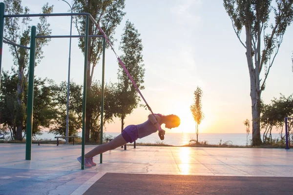 workout with suspension straps In the outdoor gym, fit woman training early in morning on the park, sunrise background