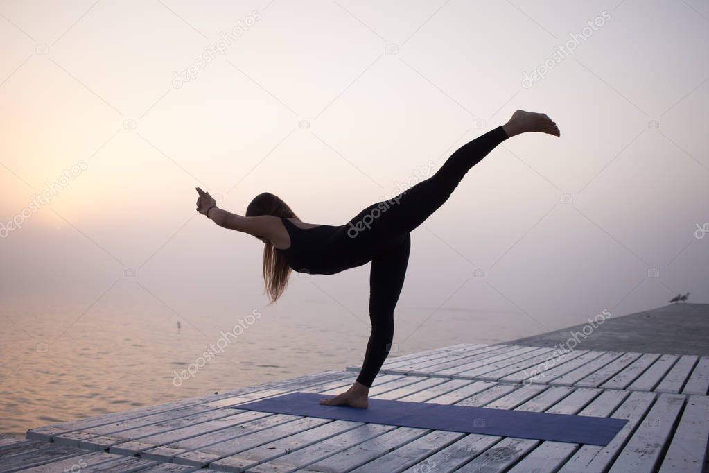 beautiful young woman in slim black wear doing yoga fitness during sunrise in fog.