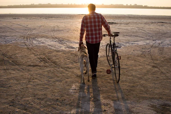 male bicycle rider walking on the sand, sunrise and lake or river background