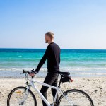 Hombre con bicicleta, hombre joven en ropa casual negro, ciclista en la playa durante el día soleado