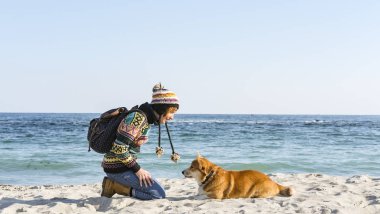 Genç ve mutlu bir kadın, sonbahar güneşli sahilde şirin bir Corgi sosisliyle yürüyor.