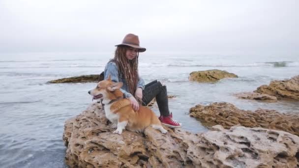 一个年轻女人带着科吉犬在海滩上摆姿势 一个女人带着恐惧的头发 一个女人带着宠物的帽子 — 图库视频影像