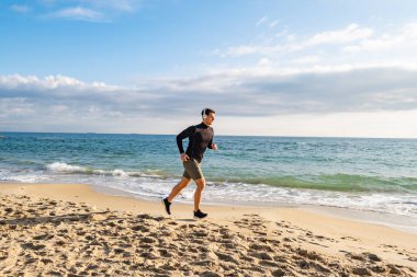 Fit male runner training on the summer beach and listen to music against beautidul sky and sea