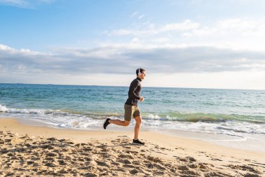 Fit male runner training on the summer beach and listen to music against beautidul sky and sea