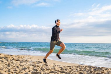 Fit male runner training on the summer beach and listen to music against beautidul sky and sea