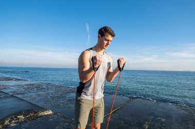 Young fit male training with  athletic rubber on the beach in summer day