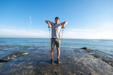Young fit male training with  athletic rubber on the beach in summer day